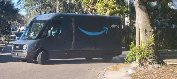 An Amazon Prime delivery van on a suburban street, showcasing the iconic blue smile logo, surrounded by trees and residential houses.