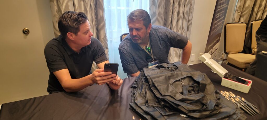 Two men at a conference table, one showing a smartphone to the other, in a discussion surrounded by conference materials