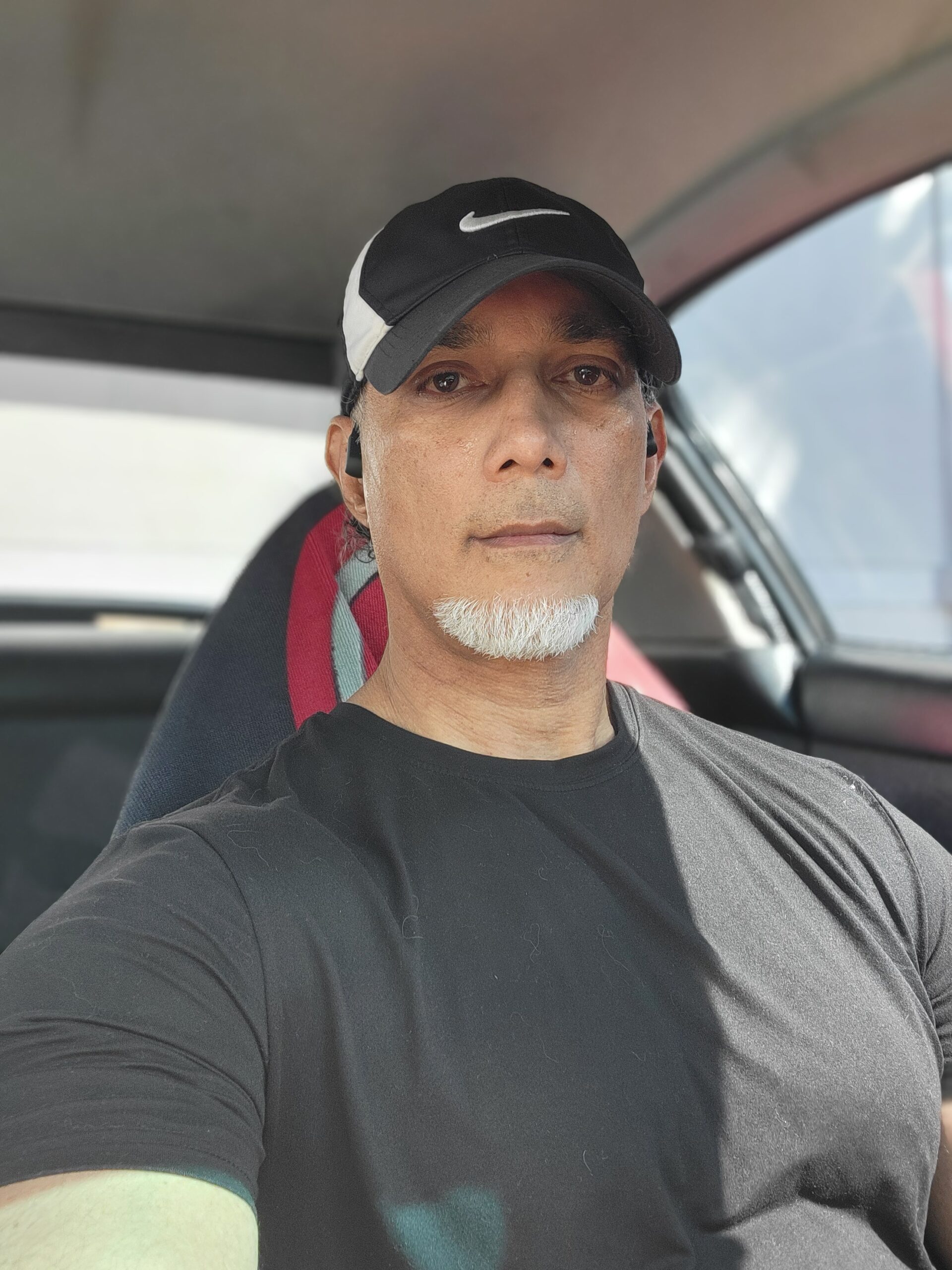 Selfie of a 58-year-old man with a white beard and mustache, wearing a black Nike cap and dark shirt, standing in front of a car, showcasing his natural body-shaping journey.