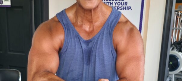 Curtis Matthews smiling at the gym after a workout, wearing a blue tank top and flexing his arms.